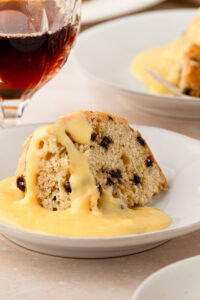A slice of classic British Spotted Dick pudding sitting in a pool of smooth yellow custard.