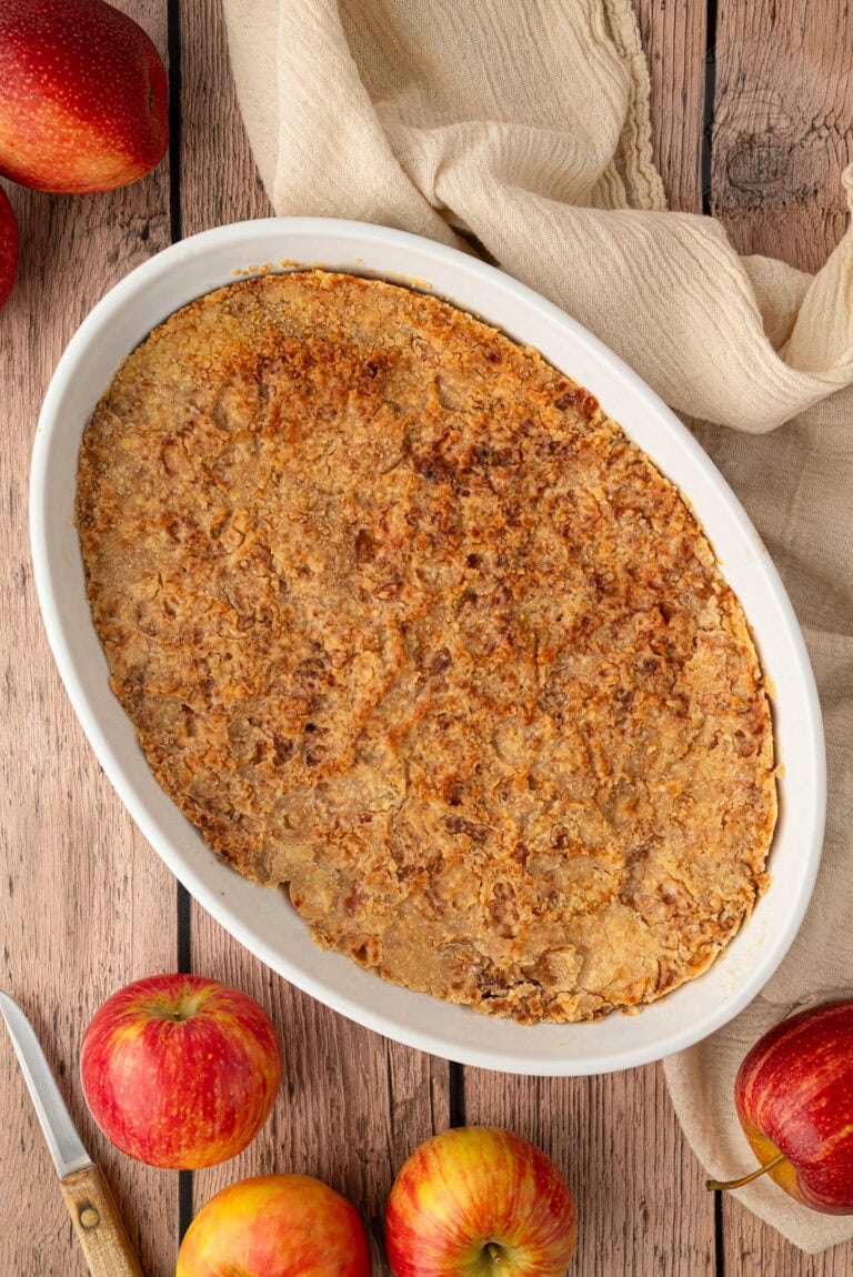 Freshly baked apple crumble in a white dish surrounded by red apples.