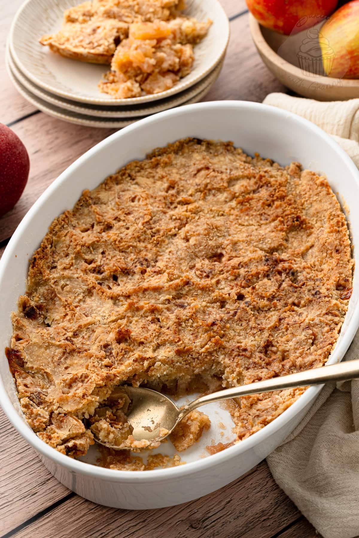Golden apple crumble in a white baking dish with a spoon serving a portion.