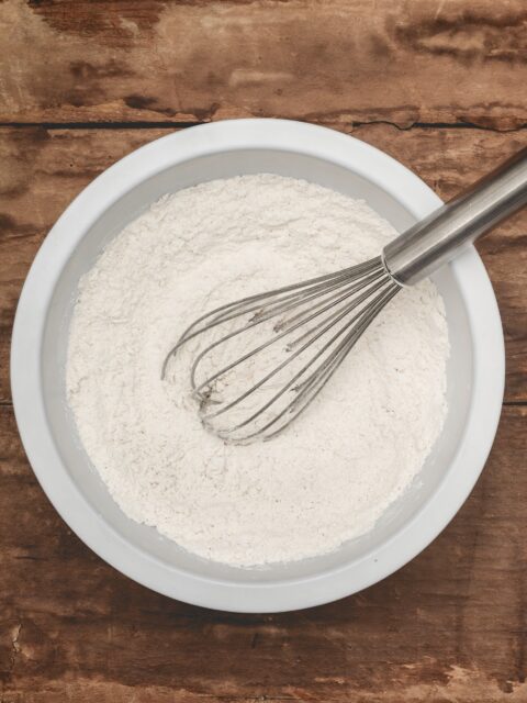 Plain flour (all-purpose flour) in a white bowl with a whisk, on top of a brown wooden surface.