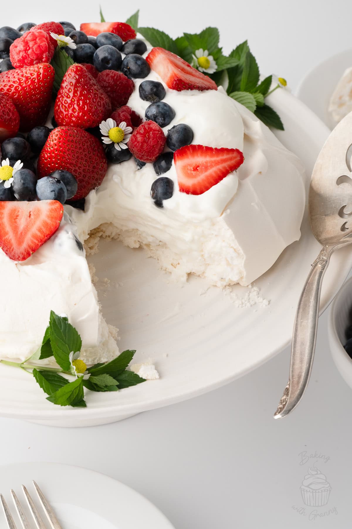 Pavlova topped with whipped cream and fresh berries, with a slice removed.