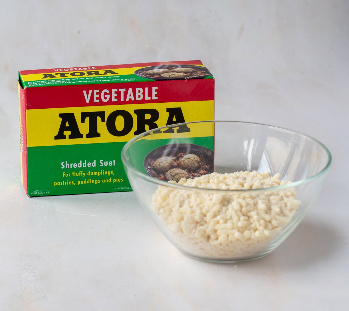 Shredded vegetable suet in a glass bowl beside a box of Atora vegetable suet.