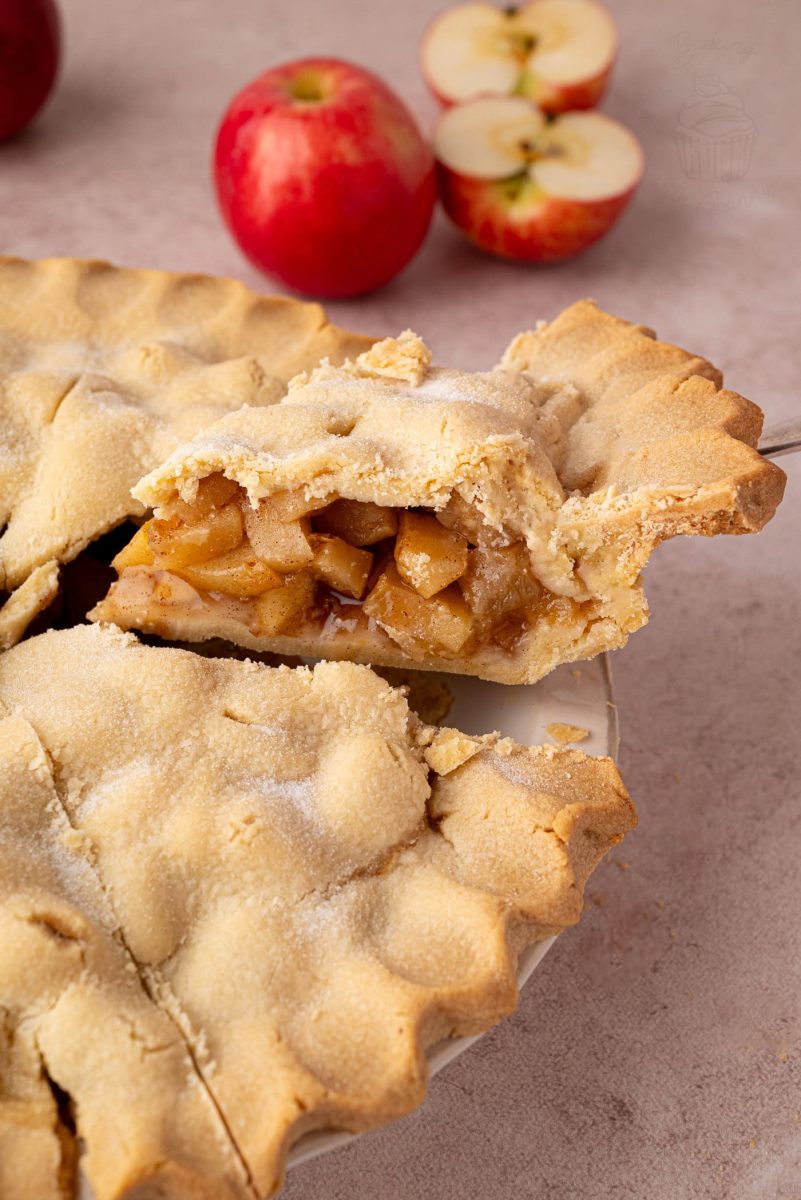 Apple Pie Slice of apple pie on a serving utensil, with whole and halved apples in the background. A traditional apple pie UK recipe ideal for autumn.