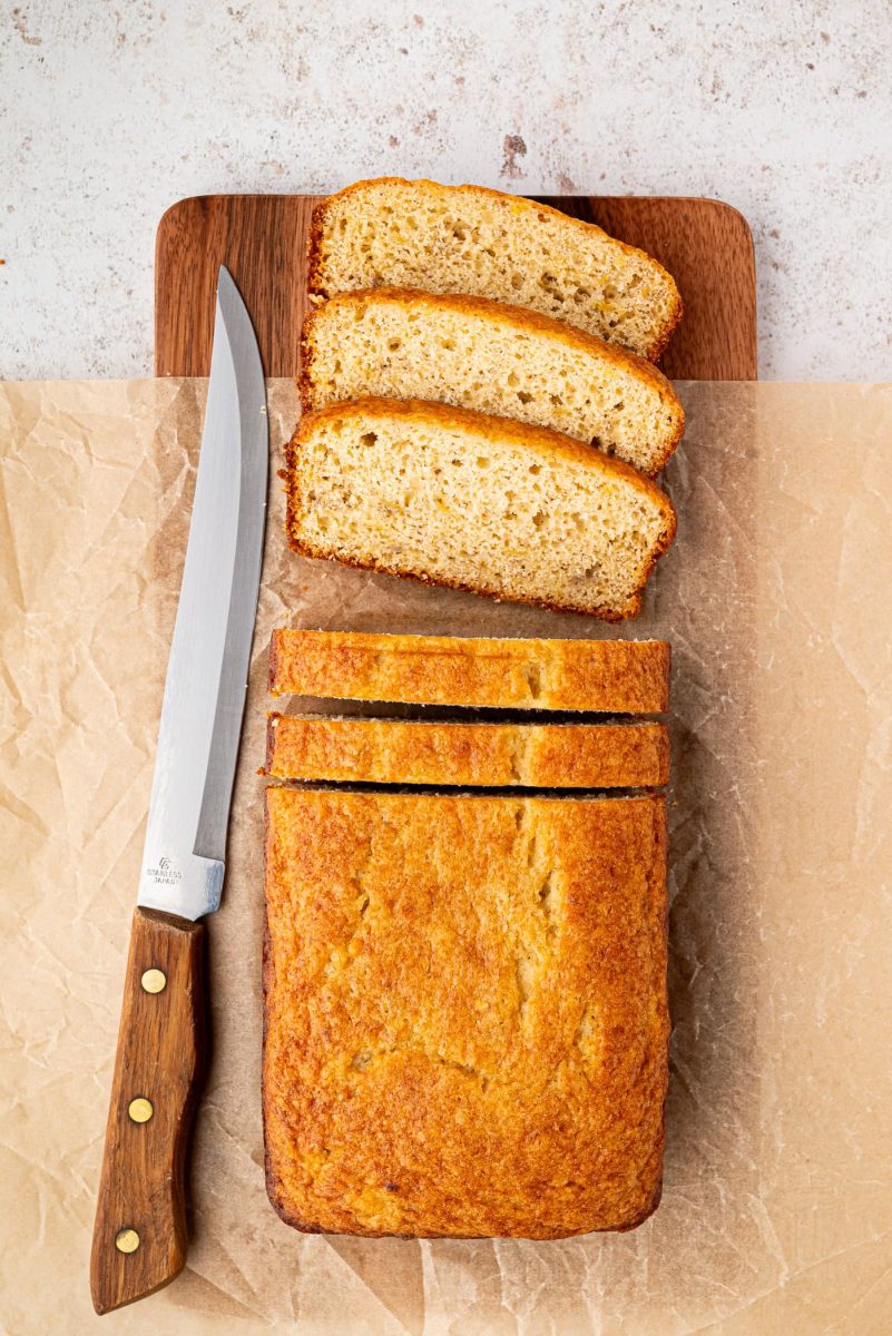 Banana Loaf A loaf of sliced banana bread rests on a wooden cutting board, with three slices arranged neatly. A knife with a wooden handle sits on the left. This inviting scene hints at a delicious banana bread recipe against the textured light background.