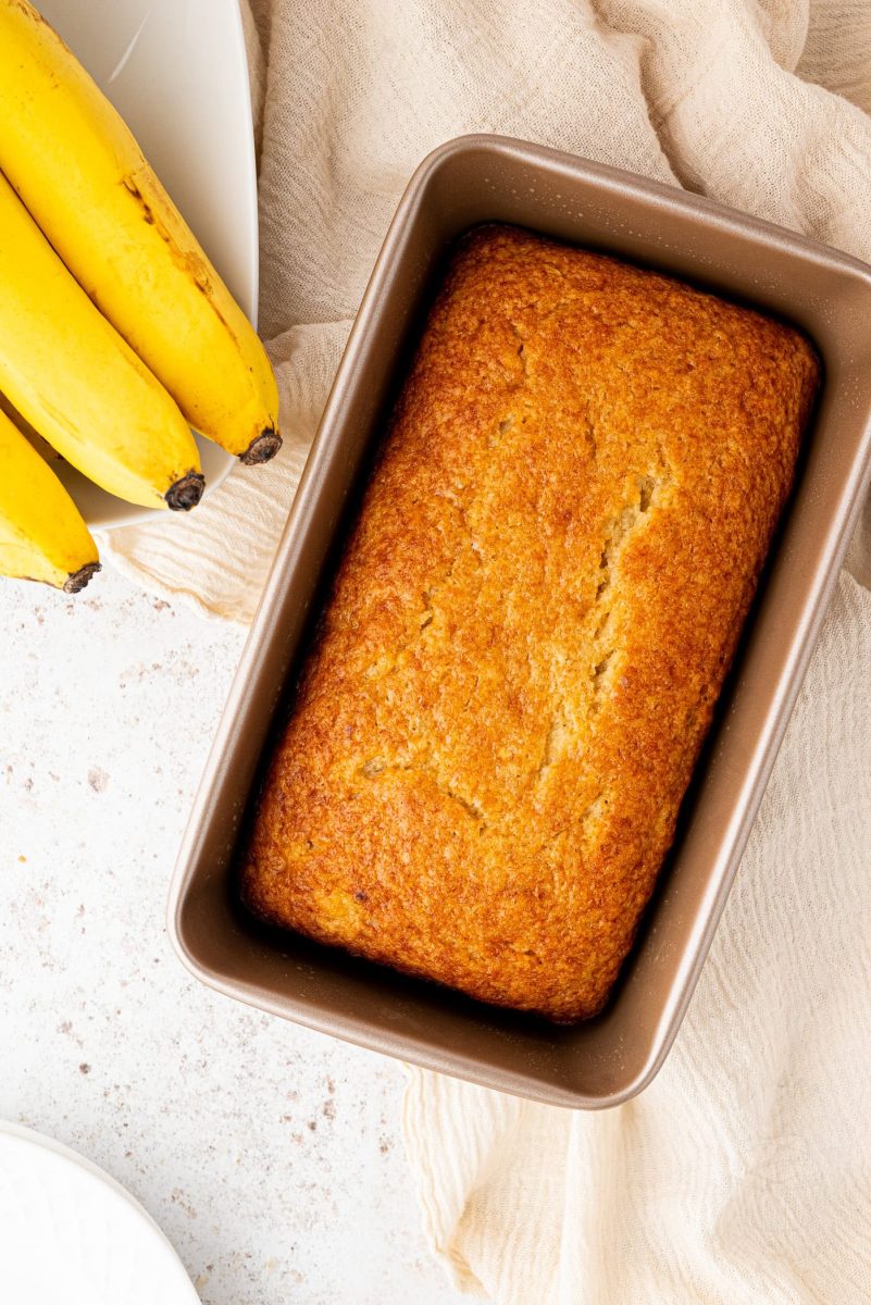 Banana Loaf A freshly baked banana loaf, made from a beloved family recipe, sits invitingly in a baking pan on the light countertop. Nearby, three ripe bananas rest on a white plate, while a beige cloth peeks out from beneath the pan.