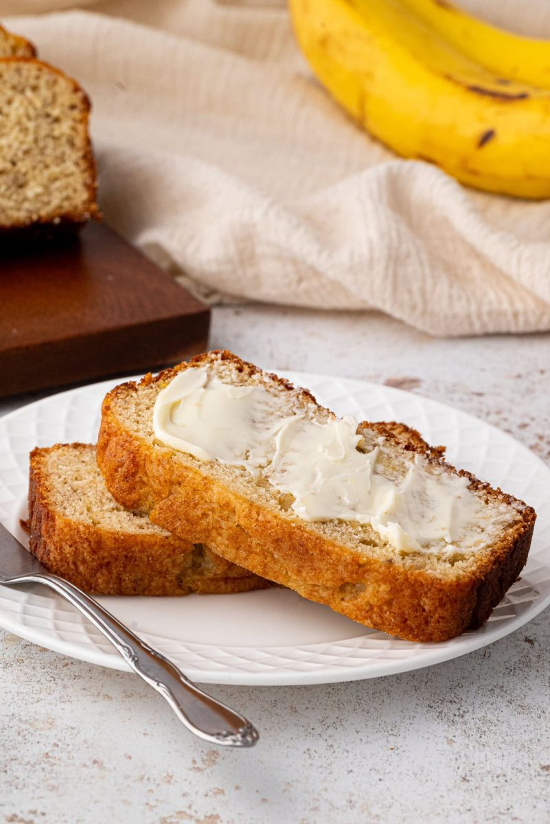 Banana Loaf Two slices of banana bread, illustrating a delectable banana bread recipe, sit on a white plate with butter spread on top. Nearby, a fork awaits indulgence. In the background, a whole banana and fresh loaf rest on a wooden board atop a beige cloth.