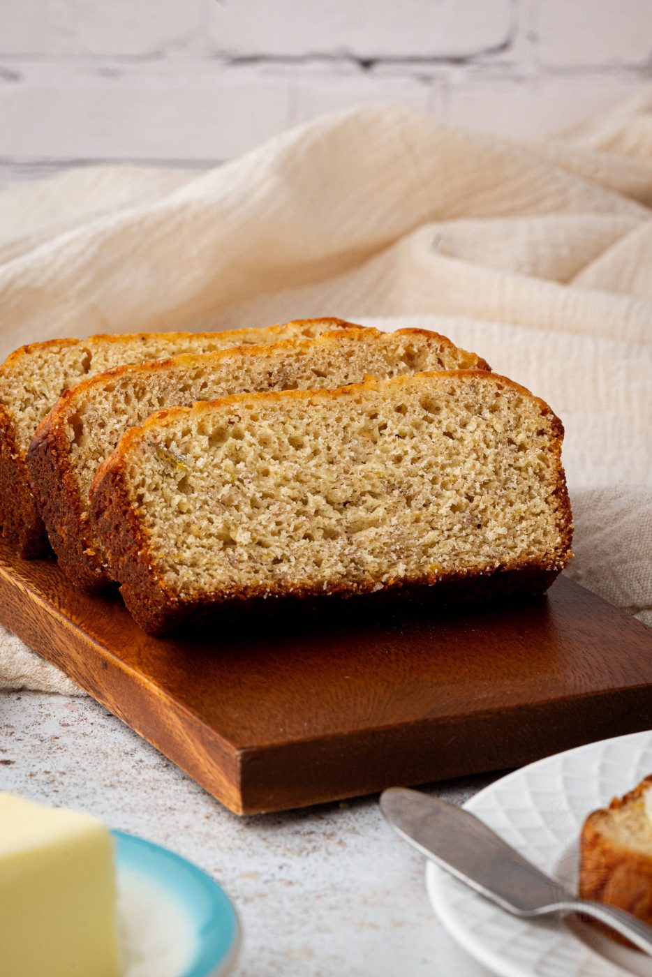 Banana Loaf Close-up of sliced banana loaf on a wooden board. The banana bread boasts a golden-brown crust and a moist, textured interior. A plate with a slab of butter and a knife is partially visible in the foreground, complemented by a cream fabric in the background.