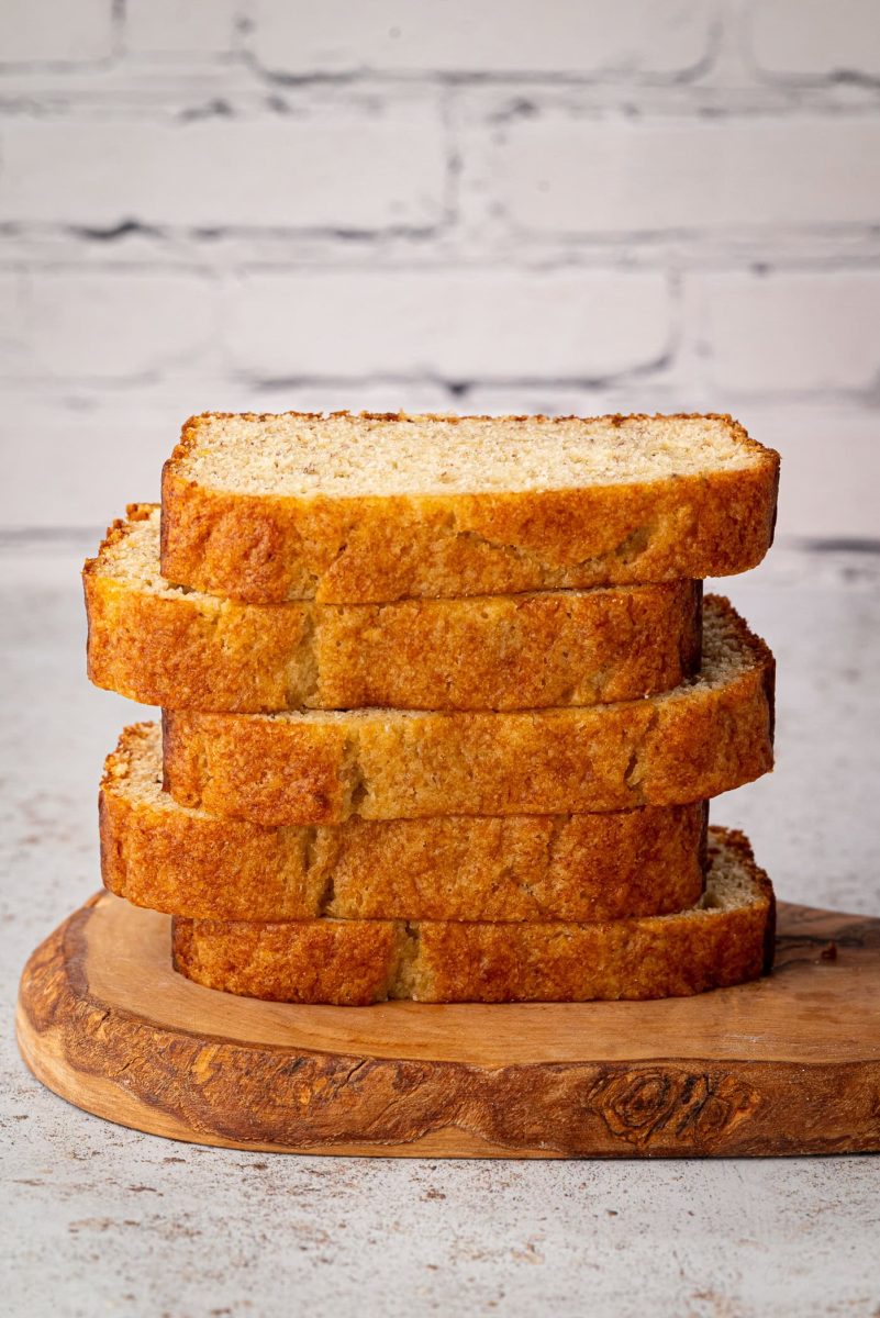 Banana Loaf A stack of five thick slices of banana loaf bread rests on a wooden cutting board, hinting at the perfect recipe. The background features a crisp white brick wall, adding charm to this cosy kitchen scene.