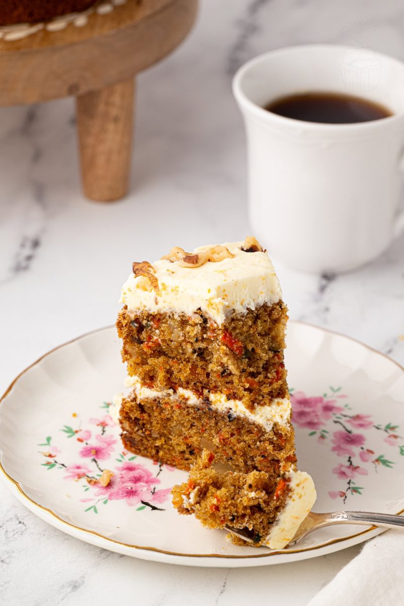 Carrot Cake A slice of moist carrot cake with buttercream frosting on a floral plate, with a bite taken on a fork and a cup of black coffee in the background.