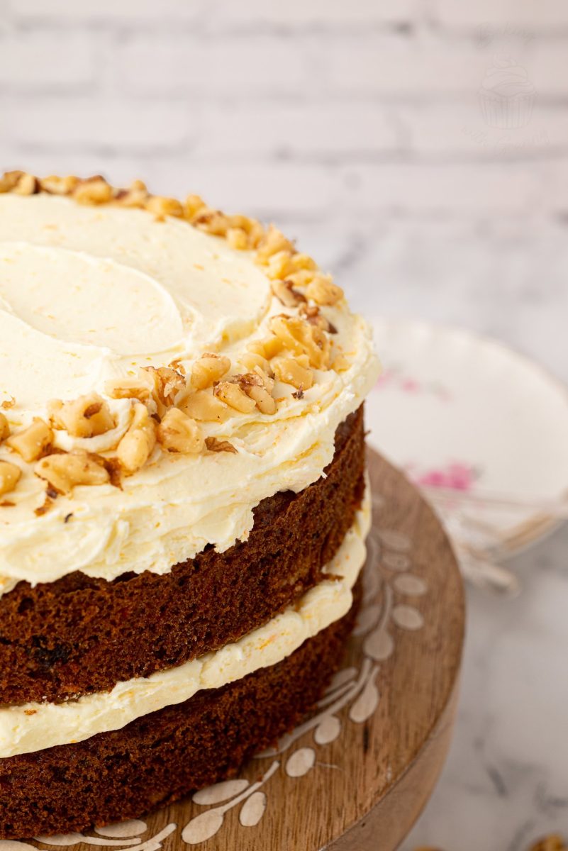 Carrot Cake Close-up of the top edge of a carrot cake, showing creamy frosting swirls and a ring of chopped walnuts on a rustic cake stand.