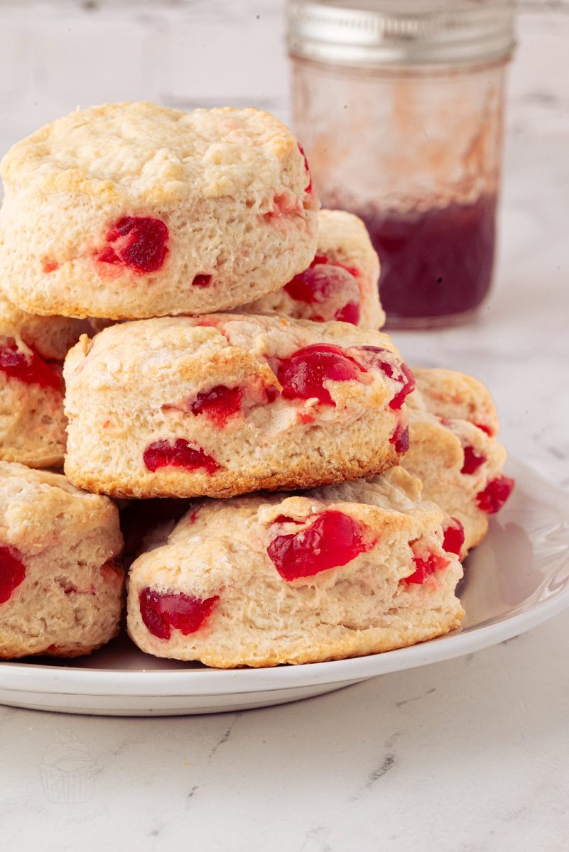 Cherry Scones A delightful cherry scone recipe comes alive with visible chunks of red glace cherries. The scones, artfully stacked on a white plate, are paired perfectly with a jar of red jam in the background on a marble surface.