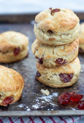 Stack of cherry scones, on a baking tray and red chequered tea towel, with other cherry scones, crumbs and glace cherries placed around.
