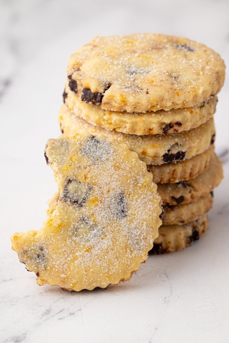 Chocolate Orange Shortbread Side view of stacked chocolate orange shortbread biscuits, with a bitten biscuit propped in front to show its crumbly texture and chocolate chunks.