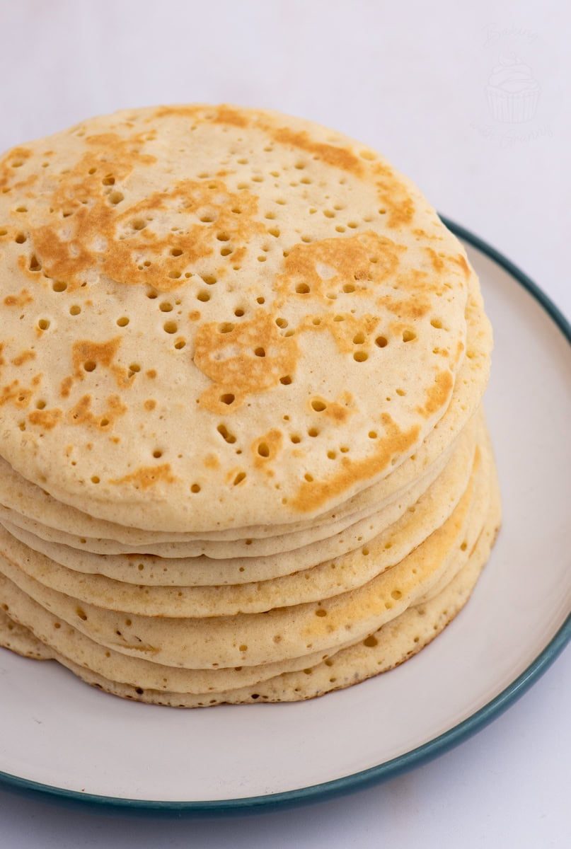 Scottish Crumpets A stack of fluffy, golden Scottish crumpets small holes on their surface, arranged on a white plate with a green rim. The crumpets are neatly stacked and appear freshly cooked against a plain background.