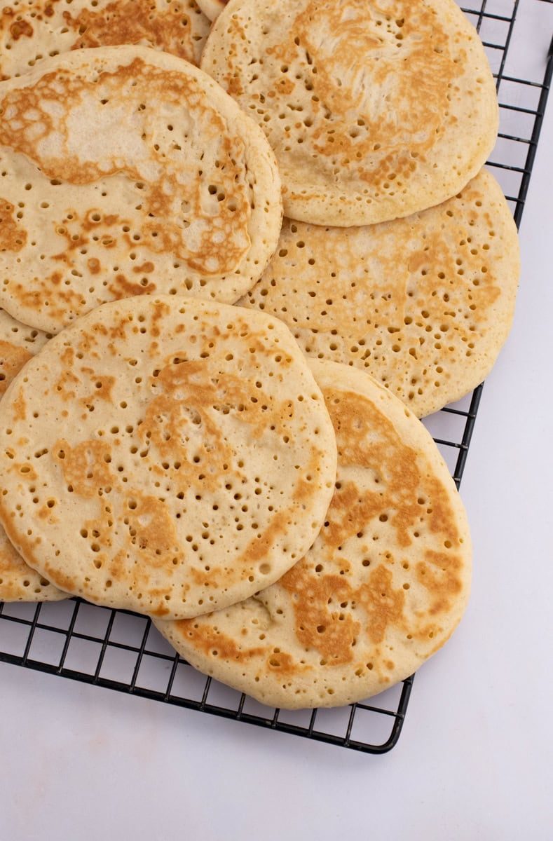 Scottish Crumpets Several round crumpets with golden-brown tops and characteristic holes, reminiscent of a traditional Scottish crumpet recipe, are cooling on a black wire rack placed on a white surface.