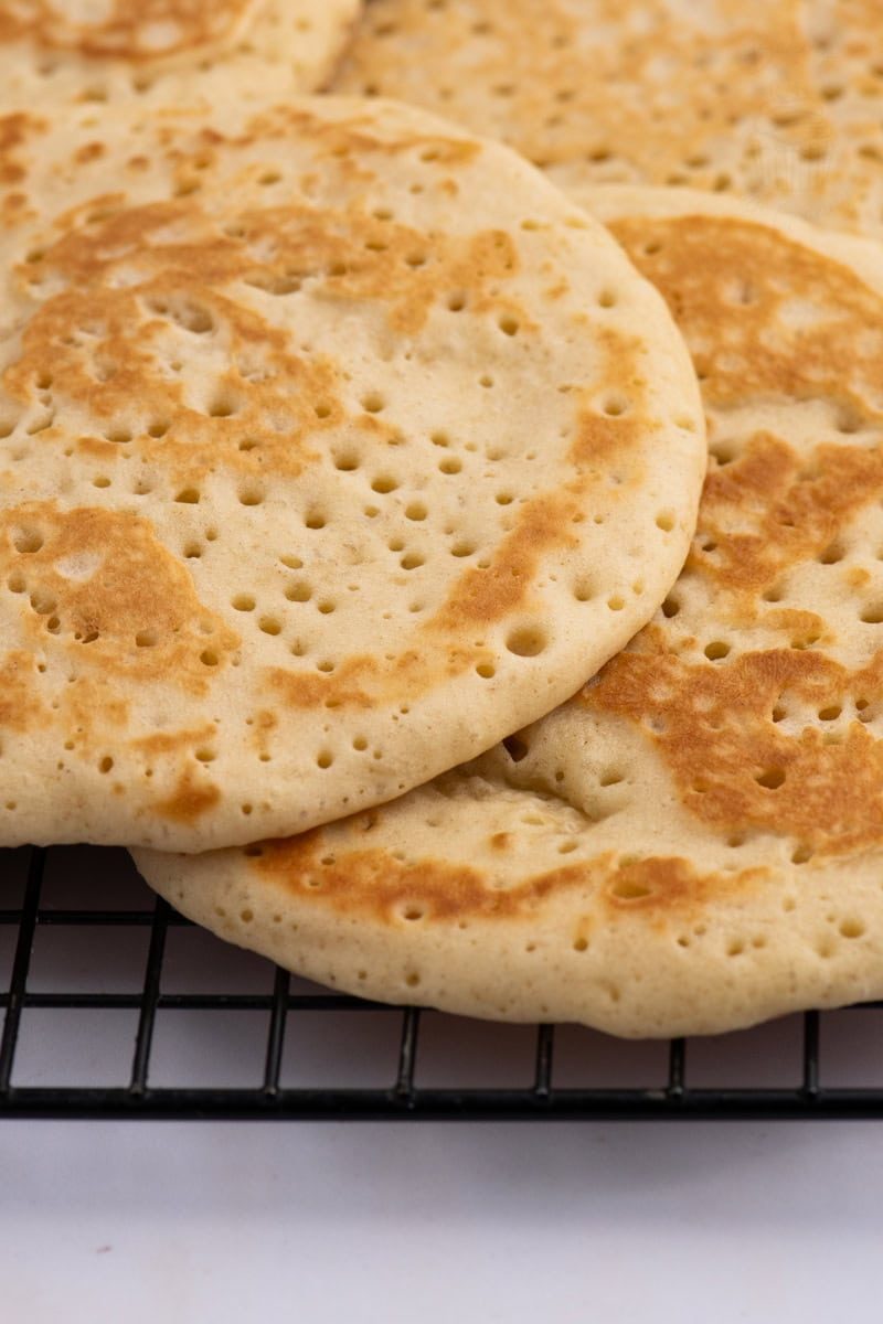 Scottish Crumpets Close-up of a stack of classic Scottish crumpets with a golden-brown surface, featuring characteristic bubbles and light texture, resting on a black wire cooling rack.