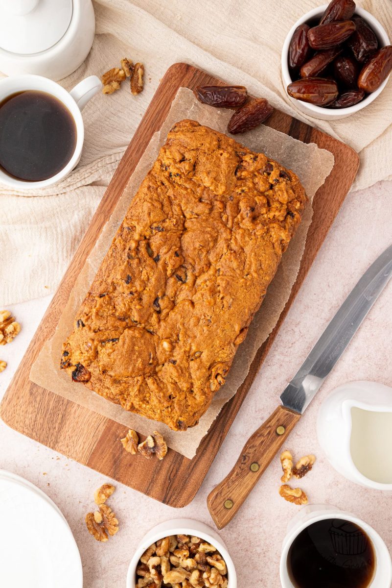 Date and Walnut Loaf A delicious date and walnut loaf sits on a wooden board, surrounded by coffee cups, dates, and walnuts. Nearby, a knife rests alongside a white milk jug and teapot on a light tablecloth.