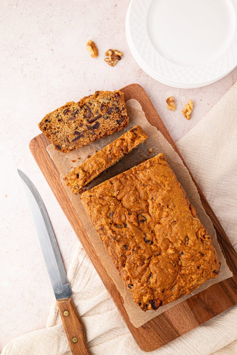 Date and Walnut Loaf A date and walnut loaf rests on a wooden cutting board. Two slices have been cut, with one placed beside the loaf. A knife lies close, while a white plate and scattered walnuts add to the inviting scene in the background.