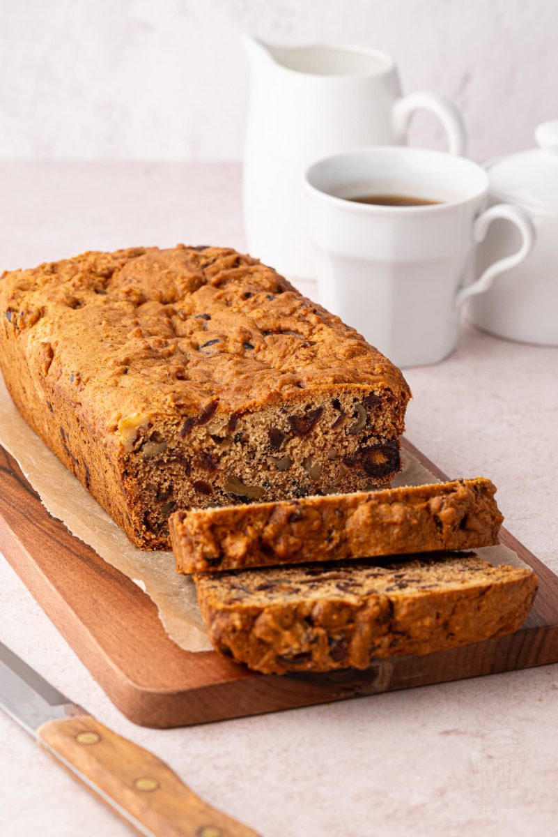 Date and Walnut Loaf A date and walnut loaf sits on a wooden board, partially sliced to reveal its rich texture with visible fruit and nuts. A knife rests nearby, while white mugs and a jug grace the light surface in the background.