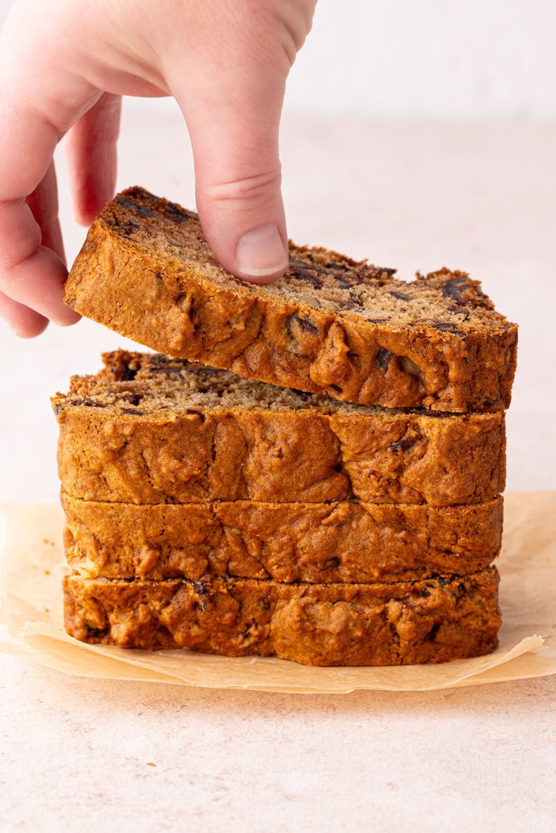 Date and Walnut Loaf A hand lifts a slice of date and walnut loaf from a stack of four on parchment paper, revealing its golden-brown crust dotted with walnut and date pieces. This delicious treat is reminiscent of a classic date and walnut loaf recipe. The softly blurred background enhances its allure.