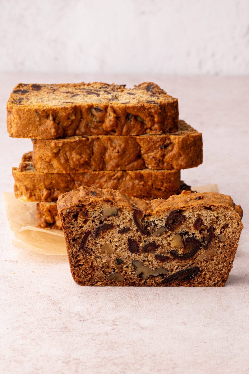 Date and Walnut Loaf A sliced date and walnut loaf reveals its moist, dense interior, adorned with visible walnuts and dates on a light surface. In the background, several golden-brown slices are stacked with parchment paper, showcasing an irresistible take on a classic recipe.