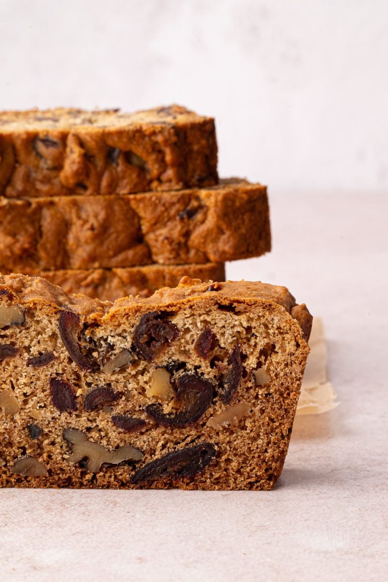 Date and Walnut Loaf Slices of date and walnut cake are stacked on a light surface. The loaf cake has a golden-brown crust with visible chunks of nuts and fruit inside, resembling a traditional date walnut loaf. The background is softly blurred, highlighting the breads texture.