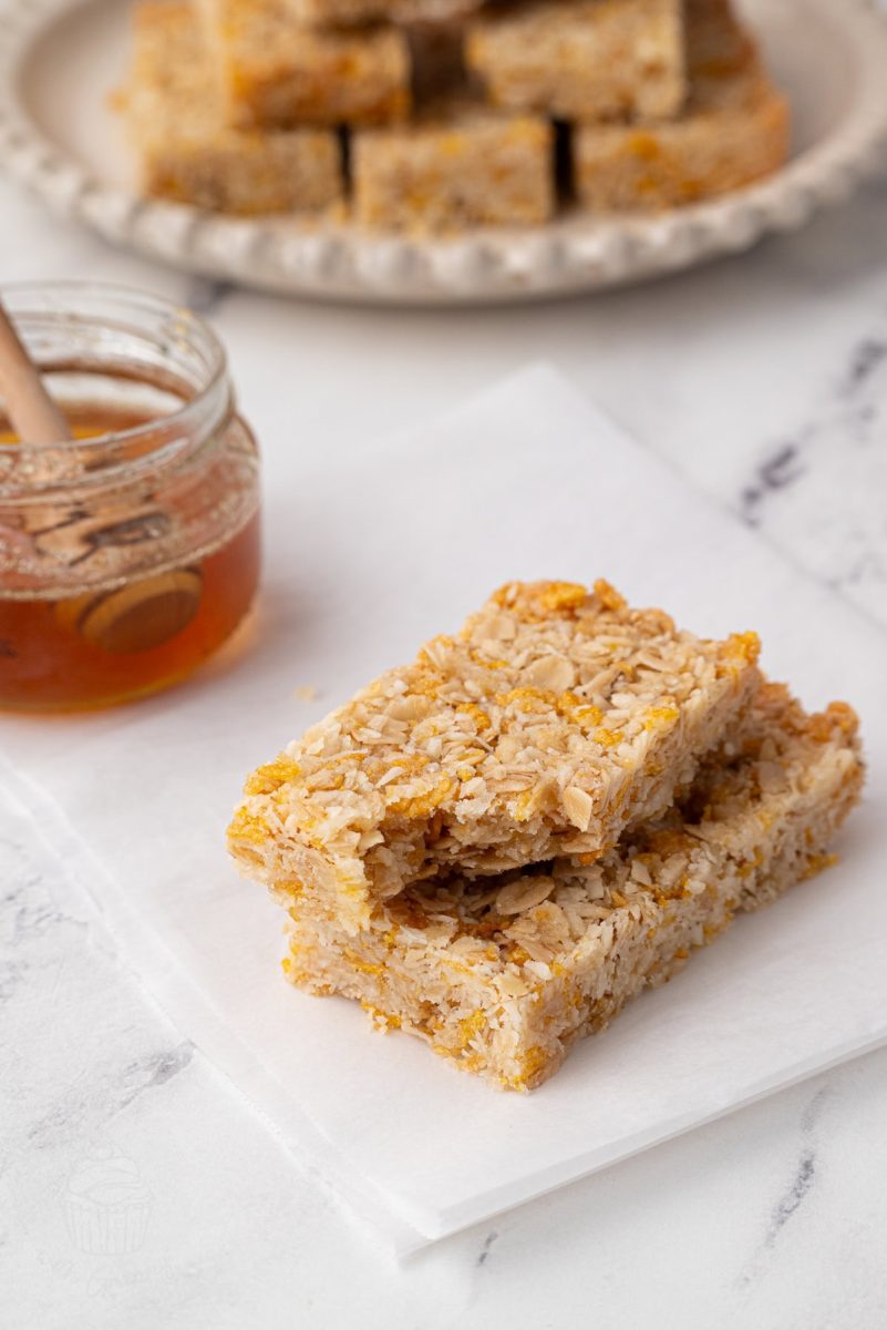 Honey Slice Two slices of honey slice stacked on white parchment paper with a jar of honey and a honey dipper in the background.