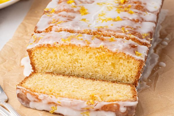 A lemon drizzle cake recipe unfolds with a lemon loaf adorned in white icing and zest, partially sliced. Resting on parchment, surrounded by fresh lemon slices and accompanied by a fork, the scene is elegantly set on a marble surface.