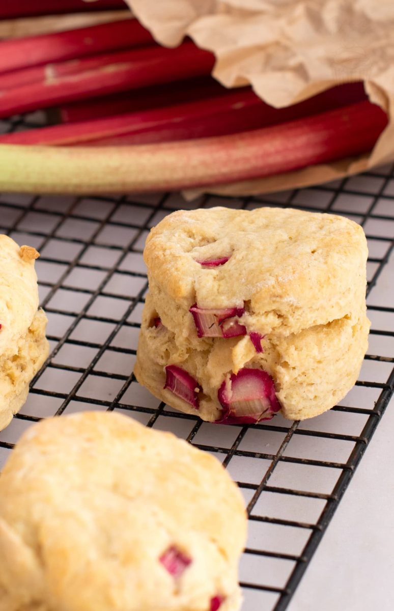 Rhubarb Scones Individual Rhubarb Scone on a cooling rack in front of fresh seasonal rhubarb stems, within a brown paper bag.