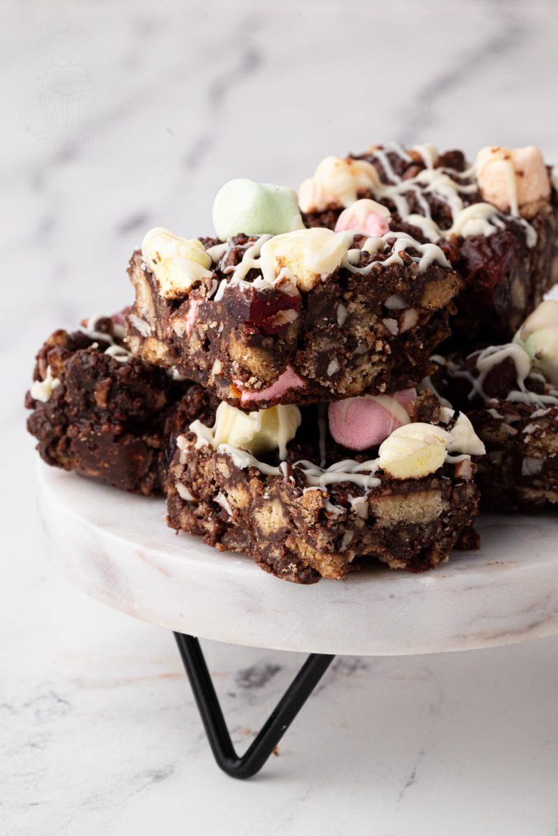 Rocky Road A stack of delectable rocky road bars sits on a marble stand, inspired by a classic rocky road recipe. These treats are made of chocolate, nuts, and colourful marshmallows, drizzled with white chocolate. The background is softly blurred against a light marble surface.