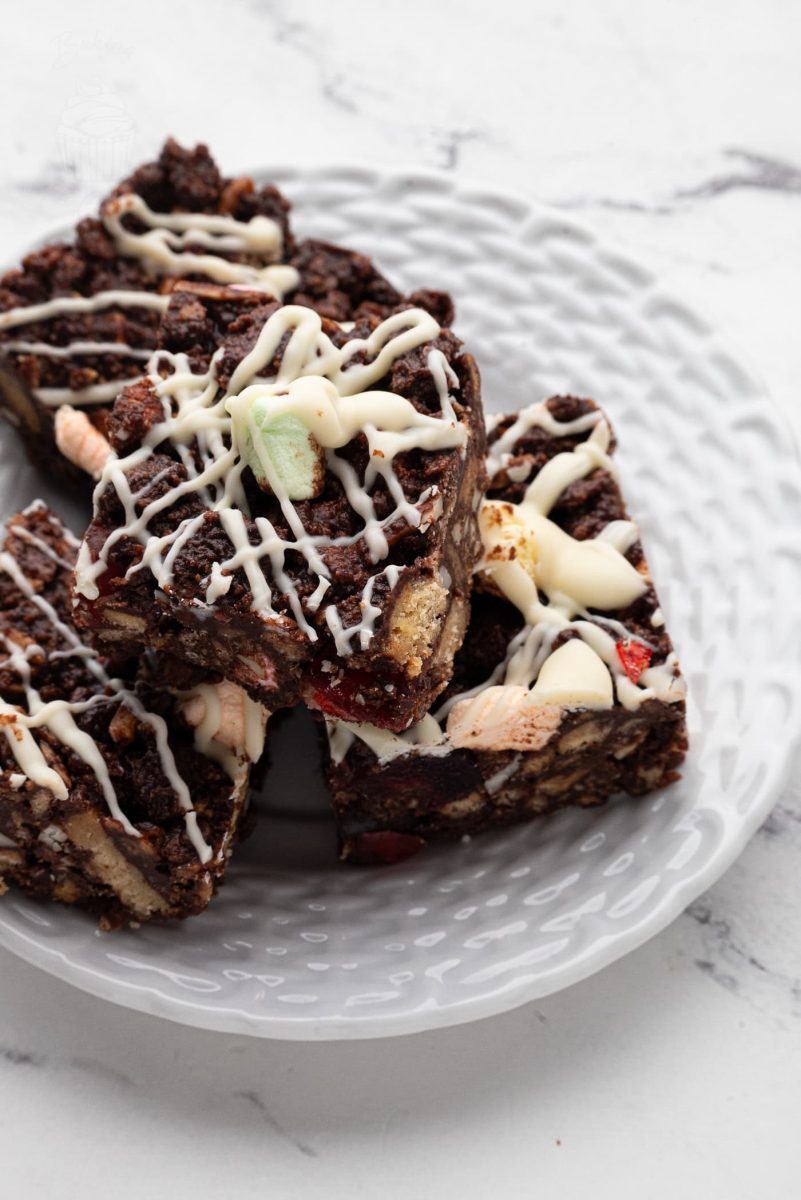 Rocky Road A white plate displays three square pieces of rocky road dessert, inspired by a classic rocky road recipe. The treats are topped with drizzles of white chocolate and contain visible chunks of marshmallows, cookies, and nuts against a white marble background.