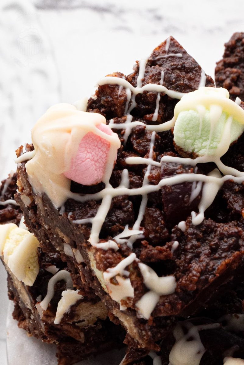 Rocky Road Close-up of stacked chocolate rocky road drizzled with white chocolate, reminiscent of a classic rocky road recipe. The brownies are topped with colourful marshmallows in shades of pink, green, and white. The dessert sits on a light marble surface.