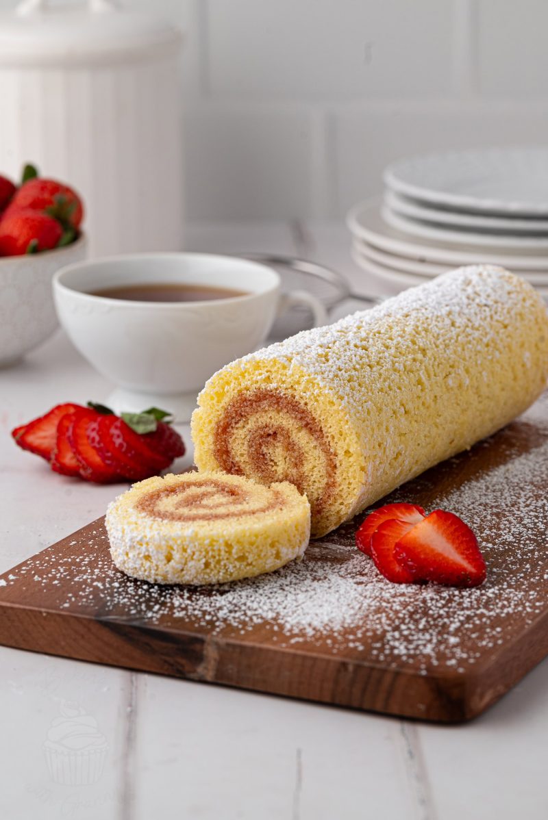 Swiss Roll A simple Swiss roll recipe is beautifully presented with a dusting of powdered sugar, partially sliced on a wooden board. Fresh strawberries are artfully arranged around it, while a steaming cup of tea and stacked plates sit invitingly in the background on a white table.