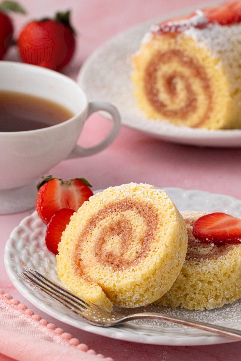 Swiss Roll A close-up of a delicious Swiss Roll cake slice, made from an easy Swiss Roll Recipe, sits on a white plate, garnished with strawberry slices. A fork rests beside it. In the background, a cup of tea and another slice of cake on a pink surface are visible.