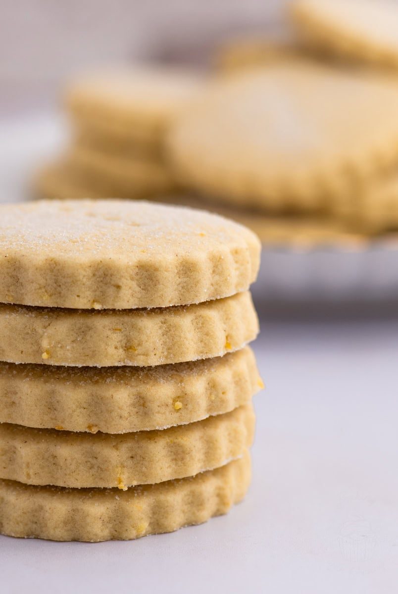 Tantallon Cakes A stack of five round Tantallon Cakes with scalloped edges is in focus. The biscuits are light golden brown with a dusting of sugar on top, reminiscent of classic shortbread. More cookies are blurred in the background on a white surface.