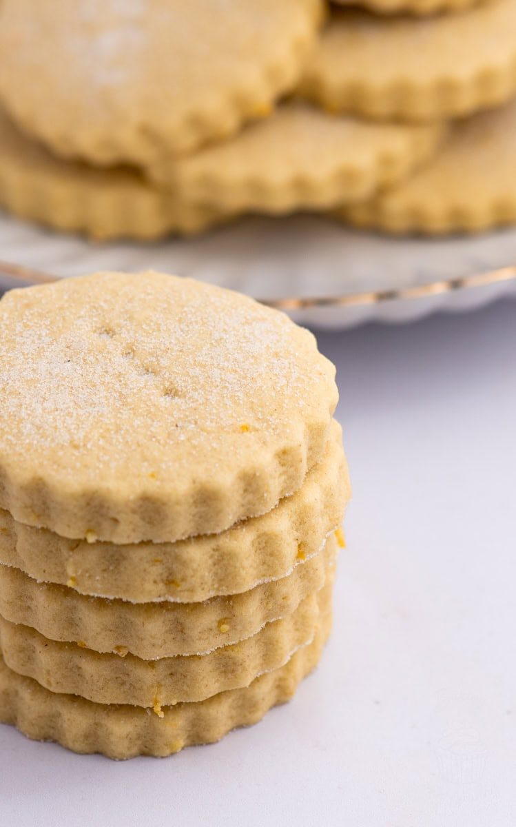 Tantallon Cakes A stack of five round, scalloped-edged, light brown biscuits with a sugar coating and specks of lemon rind, evokes the essence of a classic recipe for Tantallon Cakes. More cookies are blurred in the background on a plate. The cookies appear soft and crumbly, set against a white surface.
