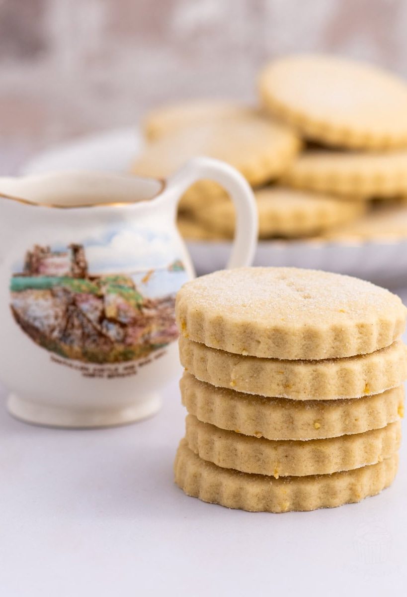 Tantallon Cakes A stack of six round, scalloped-edge Tantallon cakes sits in the foreground. Behind them is a decorative jug with a scenic illustration of Tantallon Castle in North Berwick. This delightful scene could inspire your next baking recipe, with more biscuits visible in the blurred background.
