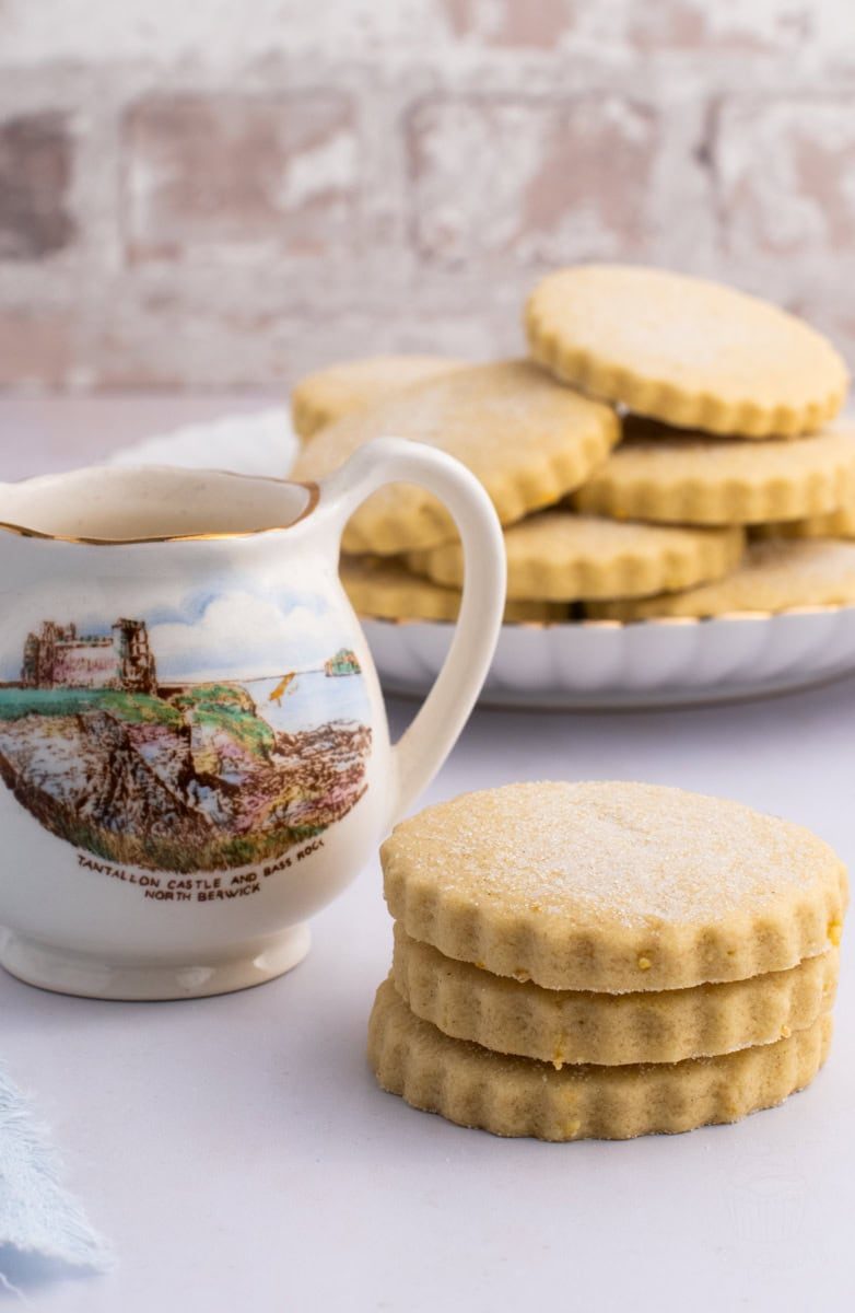 Tantallon Cakes A decorative jug with a scenic print of Tantallon Castle sits beside a stack of three sugar-dusted lemon shortbread cookies - make to the traditional recipe for Tantallon Cakes. More biscuits, are arranged on a plate in the background, all on a light-coloured surface.