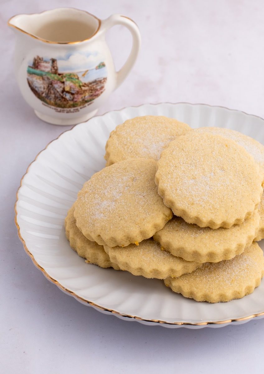 Tantallon Cakes A white plate holds several sugar-dusted shortbread cookies, reminiscent of traditional Scottish Tantallon cakes. In the background, there's a ceramic jug with a scenic painting of Tantallon Castle on it. The light-coloured surface adds a soft contrast to the delightful cookies.
