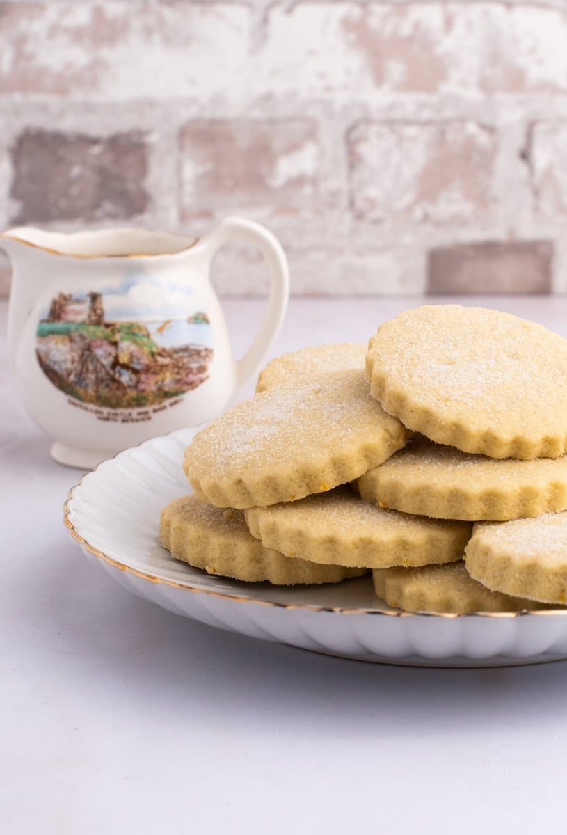 Tantallon Cakes A plate of sugar-dusted shortbread cookies reminiscent of traditional Tantallon Cakes rests elegantly on a white, scalloped-edge plate. Behind it, a decorative cream jug featuring a watercolor landscape of Tantallon Castle sits gracefully on a white surface, with a blurred brick wall in the background.