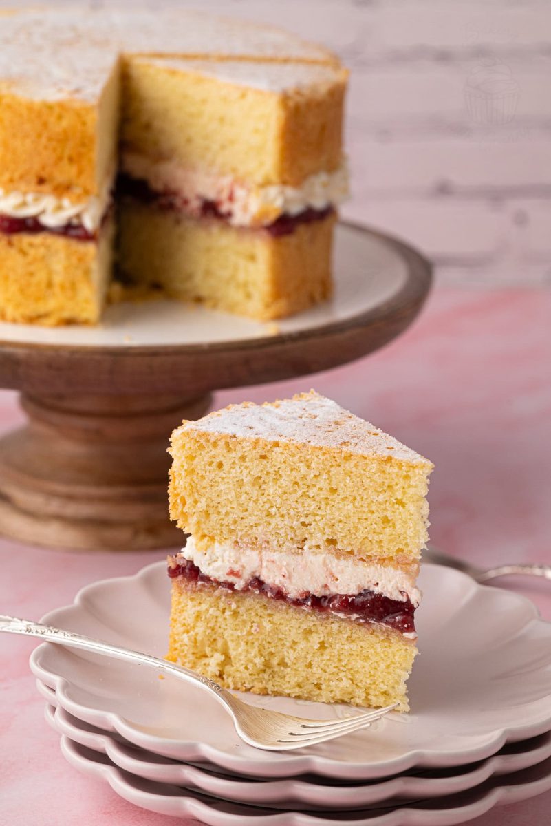 Victoria Sponge A slice of Victoria Sponge cake on a plate, showing layers of golden sponge, strawberry jam, and buttercream, with the full cake in the background.